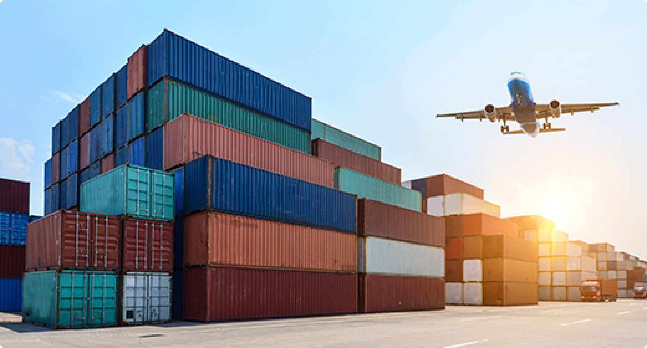 Stacked colorful shipping containers at a port with a blue cargo airplane flying overhead against a clear blue sky in the background.