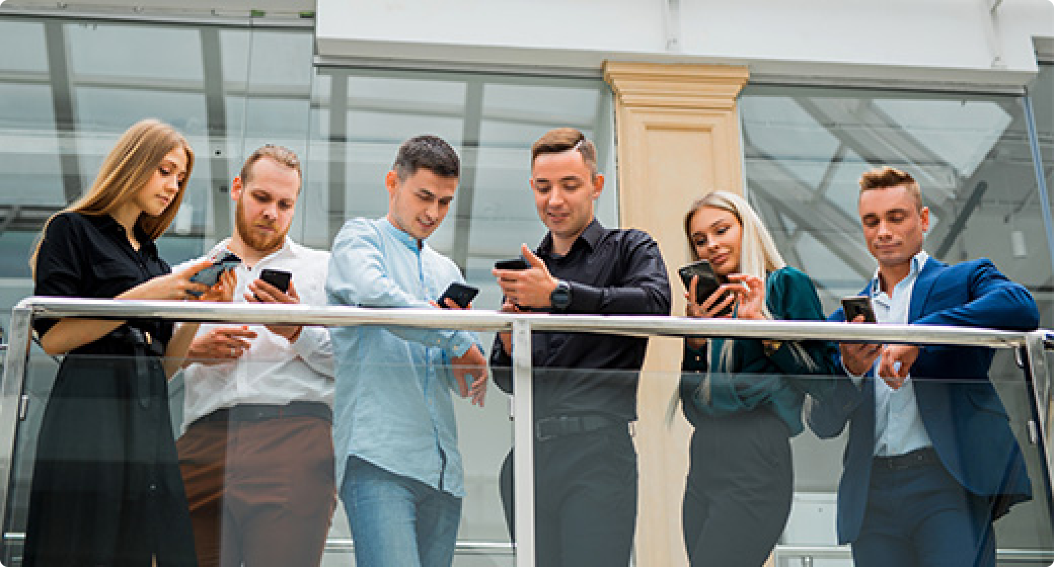 Group of business professionals standing together indoors, smiling and engaging while using their smartphones near a railing.