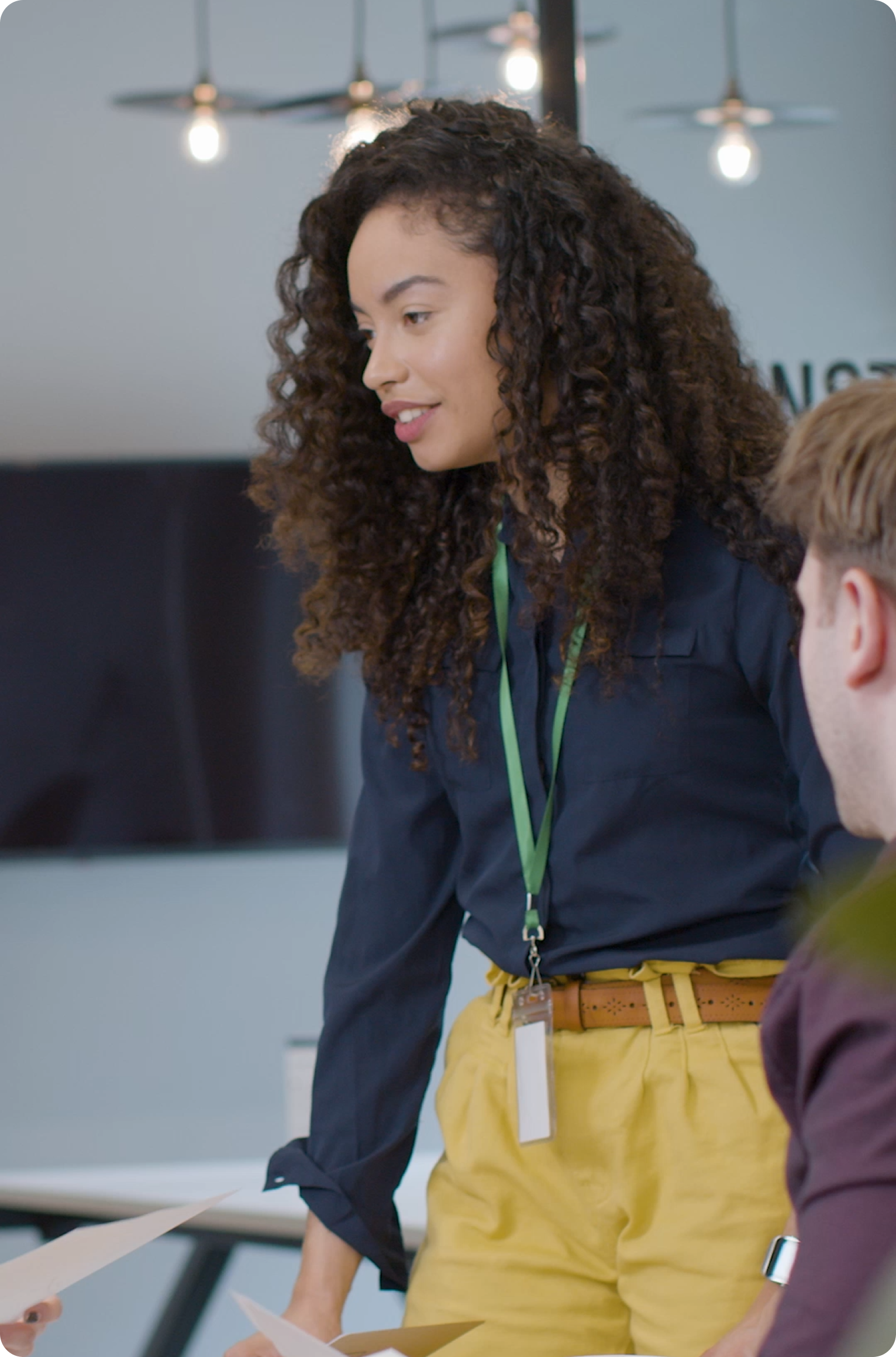Young woman in professional attire talking to colleagues in a modern office, wearing a green lanyard and standing beside a desk.