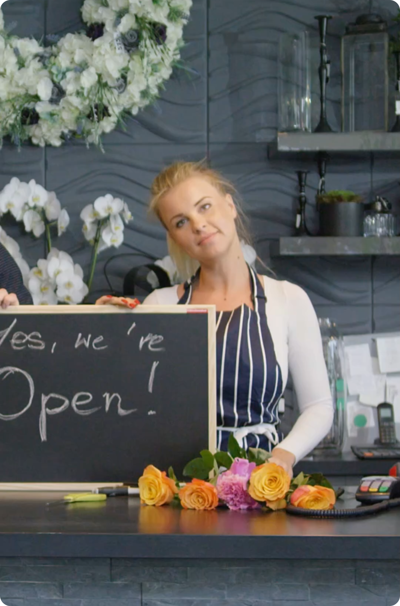 Smiling florist arranging flowers at a counter with a chalkboard sign reading ‘Yes, we’re open!’ in a flower shop decorated with white blooms.