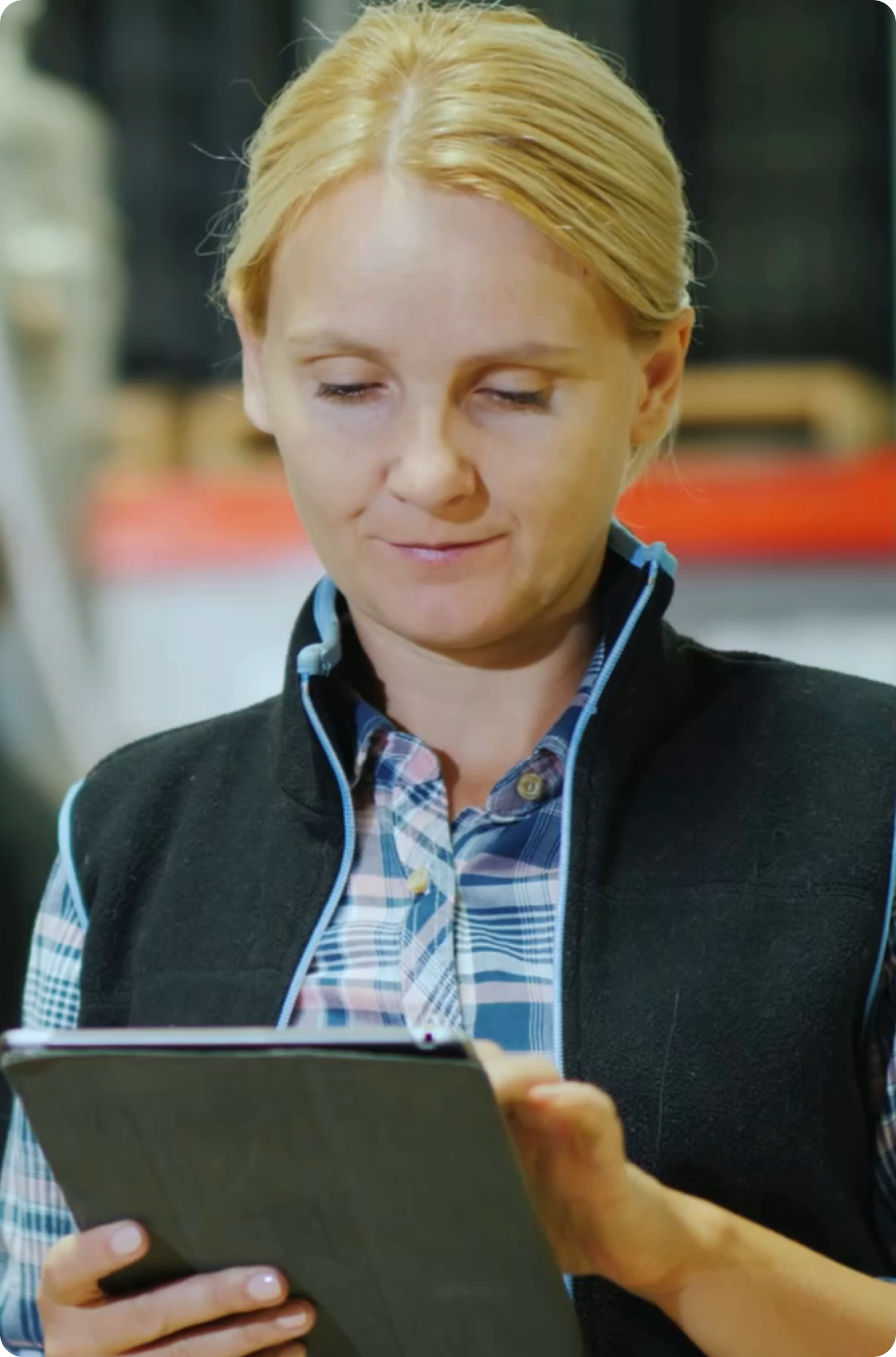 Woman wearing plaid shirt and black vest using a tablet while working in a warehouse, checking information on the device.